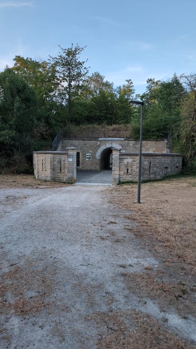 The image shows a view of an old stone fort entrance with a gravel path leading up to it. The fort has a large arched opening and is flanked by stone walls with decorative elements. There is a modern street lamp to the right of the entrance. The surrounding area is covered with dry grass and trees, suggesting an outdoor, possibly rural or park-like setting. The sky is clear blue with a few wispy clouds. There are no people visible in the image. The architecture of the fort suggests a historical military structure.