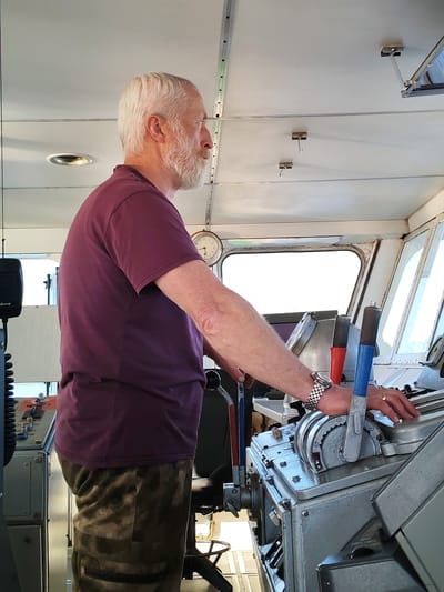 An elderly Caucasian male with white hair and a full white beard stands in profile facing right, operating a control panel on what appears to be the bridge of a vessel in Pochinok, Russia. He wears a plum-colored short-sleeved t-shirt and camouflage-patterned trousers in shades of green and brown. His left arm is bent, and his left hand, adorned with a silver-toned watch with a metal band, rests on a grey metallic control panel featuring various levers, dials, and buttons. His right hand, wearing a gold-colored ring on the ring finger, is positioned near a blue-handled lever on the main console.
The control console is constructed from grey metal, showing signs of use, with visible bolts and intricate mechanical components including a large, cog-like wheel control and red and blue plastic-handled levers. To the left of the man, a black communication speaker unit, partially labeled "Merry," is mounted on an adjacent panel. A round, analog clock displaying approximately 10:15 is visible on a panel to the man's right. The overhead ceiling is white, featuring recessed lighting, metal tracks, and suspended hooks. Large windows span the front and right side of the bridge, revealing an overexposed bright white exterior. A black swivel chair base is partially visible behind the man.
