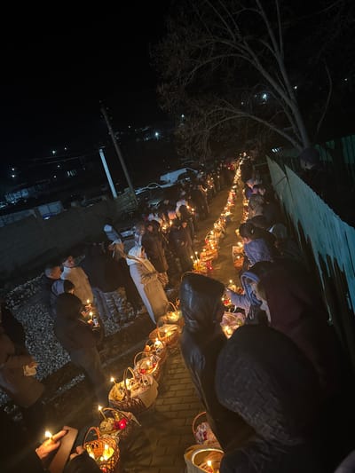 At night, a group of people stand in a line outside along a cobblestone walkway. Each person carries a lit basket decorated with lights and ornaments. Above them, bare tree branches are visible against the dark sky, and buildings are faintly illuminated in the background. The location is suggestive of a nighttime procession or festival setting. The city of Geamăna, Moldova, cannot be forensically confirmed from the image.