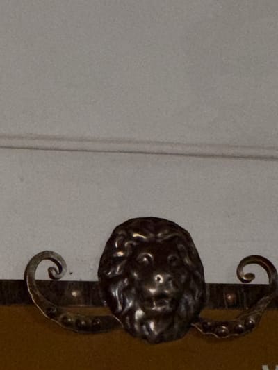 The image shows a close-up view of a decorative element on what appears to be furniture. The central focus is a metallic, sculpted lion's head, rendered in a dark, possibly aged metal. The lion's head is detailed, with visible eyes, nose, and mane. Flanking the lion's head on either side are decorative metal scrolls, also made of the same dark metal. These scrolls curve outwards and downwards, adding an ornate touch to the piece. The background is a plain, light-colored surface, possibly a wall or a painted surface, with a subtle horizontal line or seam visible across the upper portion of the image. The lower portion of the image reveals a textured, brown surface, likely the material of the furniture the decorative element is attached to. There are no people, animals, or any discernable actions taking place in the image. The location in the city of Stryi, Ukraine, cannot be determined from the visual information provided in the image.