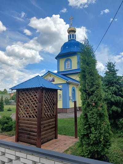 This is a low-angle, eye-level, medium shot of a church and a wooden gazebo in Doslidnytske, Ukraine. The church has a yellow facade with blue trim and a blue roof topped with a golden dome and cross. A wooden gazebo with a blue roof is located in front of the church. A tall, green coniferous tree stands to the right of the gazebo, partially obscuring the church. In the background, there are more trees and a few houses. The sky is blue with white, fluffy clouds. The ground is covered with green grass and a paved walkway. A brick wall is in the foreground. There are no people or animals visible in the image.