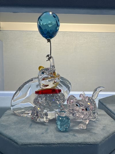 The image shows a crystal Dumbo figurine holding a blue ball in its trunk, with a crystal Timothy Mouse figurine beside it. The objects are displayed on a blue, padded surface, likely a display case. The background includes a white wall or panel with a visible edge suggesting a corner. The location is Spreitenbach, Switzerland, based on the Dumbo figurine being available at the Swarovski store in the Shoppi Tivoli shopping center in that city.