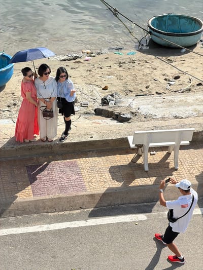This media file presents an outdoor scene in Ninh Chu Ward, Vietnam, under bright, clear conditions, evidenced by strong shadows. The image is originally rotated 90 degrees counter-clockwise; when viewed upright, the scene depicts a waterfront area with people engaging in leisure activities.

In the foreground, three women are standing on a sandy and somewhat muddy bank adjacent to the water. One woman, wearing a light blue striped blouse and a black skirt, gives a thumbs-up gesture. Next to her, another woman in a light-colored top with lace details and long cream-colored pants holds a small brown purse. The third woman, dressed in a coral-orange dress, holds a blue umbrella, presumably for shade. They appear to be posing, perhaps for a photograph. The bank itself shows some scattered debris.

Partially submerged in the water and tied to the bank is a light blue, traditional Vietnamese basket boat (thúng chai).

Above the women, on a paved sidewalk with decorative patterned tiles and an adjacent asphalt road, a young man is visible. He is leaning forward, holding a smartphone in his hands, seemingly taking a photo or looking at content on the device. He wears a white baseball cap, a white t-shirt with red stripes on the shoulder, black shorts, and red and black athletic shoes. A black fanny pack is worn across his chest, and he has a watch on his left wrist.

Between the paved area and the sandy bank, a simple white concrete table or bench can be seen. The time of day appears to be mid-morning or early afternoon due to the high sun angle and distinct shadows. The overall atmosphere is relaxed, with individuals enjoying the sunny weather by the water. No distinct text is clearly legible on any objects or clothing.