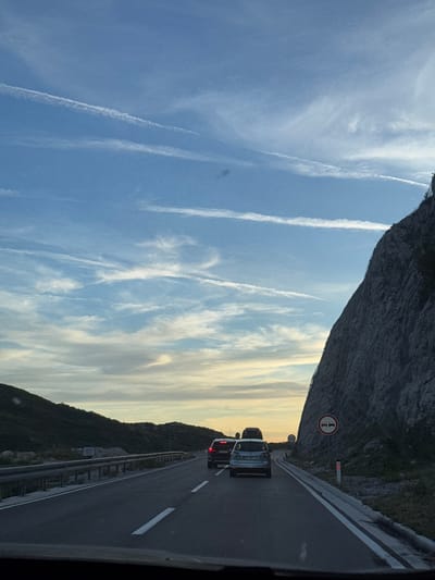 The image depicts a highway scene with vehicles driving along the road. The highway is flanked by a steep, rocky mountain on the right and a grassy embankment and smaller hills on the left. The sky is a clear blue with scattered white contrails from airplanes and wispy clouds, suggesting it might be late afternoon or early evening. There are several cars on the road, including a silver hatchback in the foreground and a dark SUV behind it. A traffic sign indicating "no overtaking" with two cars in a red circle is visible on the right side of the road, near the mountain. Another sign with "60" is also present. The overall environment appears to be a mountainous coastal region, consistent with the area around Obzovica, Montenegro, which is known for its scenic coastal highways. The lighting suggests a time of day with softer sunlight.