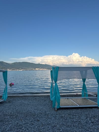 The image depicts a serene beach scene in Neberdzhaevskaya, Russia. In the foreground, a gravel beach is occupied by a white canopy with turquoise curtains, suggesting a private lounging area. The canopy has a wooden platform for seating or reclining. In the middle ground, the calm blue sea gently laps against the shore. A person is visible in the water, wearing a pink inflatable ring and seemingly enjoying a swim. In the background, a picturesque mountain range forms a majestic backdrop to the coastal town, which is dotted with buildings and structures. The sky above is a clear, vibrant blue, with large, fluffy white clouds adding depth and texture to the horizon.