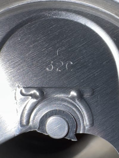 The image displays a detailed, extreme close-up of the interior surface of an aluminum beverage can lid, reportedly located in Peschiera Borromeo, Italy. The surface is primarily metallic silver-grey with a distinct brushed texture, showing parallel striations and reflections from an overhead light source. In the upper central portion, the alphanumeric sequence "32C" is debossed into the metal. Below this marking, the structural components of a pull-tab opening mechanism are visible. This includes a circular rivet head in the lower center, securing the (unseen) pull-tab, surrounded by concentric, raised, and depressed metal contours that form part of the lid's score line and opening mechanism. The edges of the formed metal exhibit minor irregularities. No individuals are present, nor are there any discernible environmental features to confirm the stated geographical location.