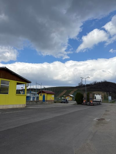 The image depicts an outdoor scene at what appears to be a facility entrance or parking area in Gorna Rositsa, Bulgaria, under a partially cloudy sky with prominent white and gray clouds and visible blue sky.
In the left foreground is a single-story, bright yellow building with a brown wooden roof trim. It features a window with horizontal security bars and an open service counter/hatch, on which a decorative figurine is placed. A small white satellite dish is affixed to the building's exterior.
Behind this yellow building, a blue and white structure is visible, bearing signage that reads "PRESS CENTER" and "SEVLIEVO THE BEST." To its right, another smaller yellow building with a red roof and a blue door displays partial signage, including the word "TRAK." A Bulgarian flag is visible in the background, between the first yellow building and the blue structure.
In the middle ground, a dark-colored SUV, consistent with a Jeep Wrangler model, is parked. One individual is standing next to the driver's side of this vehicle. Further to the right, a white semi-truck tractor unit is visible, pulling an unladen low-loader trailer equipped with ramps and red-and-white reflective markings on its rear.
The ground is predominantly dark asphalt pavement, featuring visible manhole covers. Multiple tall streetlights are positioned throughout the scene. The background consists of gently sloping green hills, some showing areas of disturbed earth or bare ground consistent with a track, and sparse deciduous trees.