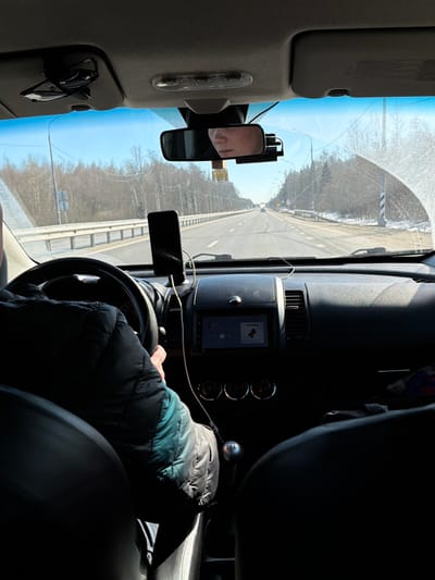 The image depicts the interior of a moving vehicle from a rear passenger's perspective, looking forward through the windshield onto a road in Spas-Zaulok, Russia. A driver is visible on the left, wearing a dark and teal puffer jacket, with their left hand gripping a black steering wheel. A manual transmission gear stick with a silver knob is visible on the center console. A black smartphone is mounted vertically on the dashboard and connected by a white cable. The vehicle's infotainment screen displays a music interface with the text "МУЗЫКА" and an album art icon. Above the windshield, dark folded sunglasses are clipped to the overhead console, and a rearview mirror reflects the lower half of a person's face. A black dashcam is mounted to the right of the rearview mirror, and a small, rectangular air freshener hangs from the mirror stem. The windshield is noticeably dirty or smudged, particularly on the right side.
Outside the vehicle, a multi-lane asphalt road with white markings extends into the distance under a clear blue sky. White metal guardrails line both sides of the road. The surrounding environment features bare trees, utility poles, and patches of snow on the ground along the roadside, suggesting a late winter or early spring setting. Distant vehicles are visible on the road ahead.
