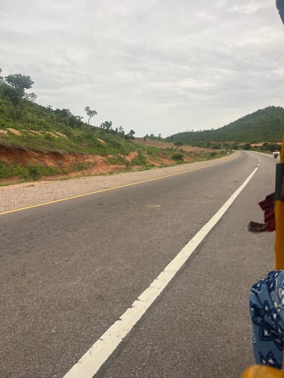 The image captures a view from a vehicle, likely a tricycle given the yellow framing visible at the top. The road ahead curves gently to the left, bordered by a white line on the right and a yellow line on the left. On the left side of the road, a lush green, tree-covered hillside rises steeply. The hillside is interspersed with patches of exposed red earth and rocks, suggesting a natural, somewhat rugged terrain. A few scattered trees stand out against the dense greenery. The sky is overcast with a layer of pale gray clouds, indicating a likely dull or rainy day. The overall mood is one of travel through a natural landscape. The scene appears to be a roadside view, with no specific landmarks that would pinpoint an exact location within Minda, Nigeria, beyond it being a road traversing a hilly, vegetated area. The time of day is indeterminate due to the overcast sky, but it appears to be daytime. There is no visible text in the image.