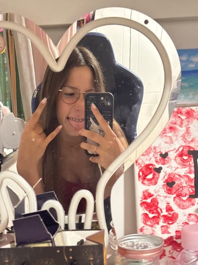 This media file is a photograph taken in Prolesha, Bulgaria. It shows a young woman with braces and glasses, taking a selfie in a heart-shaped mirror. She is wearing a red top and has her hair down. Her nails are painted black and white. She is holding a smartphone and making a peace sign with her fingers. In the background, there is a painting with red poppy flowers. Various objects are visible around the mirror, including a pink object and a metallic container. The setting appears to be indoors, possibly a bedroom or a vanity area. The lighting suggests it is daytime. The woman's expression seems playful and confident.