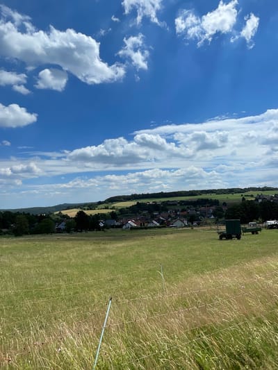The image depicts an outdoor scene, likely in Schwarzenborn, Germany. The composition is dominated by a vast blue sky filled with scattered white clouds, suggesting a fair weather day. A narrow band of trees borders a village with houses, indicating a rural or semi-rural setting. The foreground consists of a grassy field, partially enclosed by a fence or barrier made of thin wires supported by stakes.

A dark green structure, possibly a shed or a small building, is visible in the middle ground, situated near a vehicle that appears to be a trailer or a farm implement. The field itself seems to be a grassy area, possibly a park, a meadow, or a sports field, with some dry grasses mixed in. A light blue pole extends diagonally across the fence, suggesting it might be part of a temporary structure or a marker.

The overall impression is that of a tranquil summer day. The lighting suggests it is daytime, likely afternoon, given the brightness and the angle of the shadows (though shadows are not very prominent in this particular orientation). There is no visible text in the image. The scene conveys a sense of peacefulness and openness.