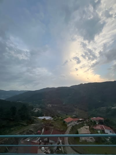 The image shows a view in Amarante, Portugal, captured from a high vantage point, possibly a bridge or elevated structure. The scene includes a mountainous landscape with homes situated on the hillside. The sky is cloudy, with a patch of sunlight breaking through. Some blue railings are visible, indicating the viewpoint’s structure.