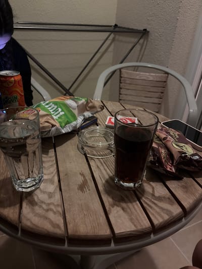 This image captures a close-up view of a small, round, slatted wooden table located on an outdoor patio or balcony in Selçuk, Turkey. The table is adorned with several items indicative of a casual gathering.
On the left side of the table, a clear glass filled with sparkling water, exhibiting numerous small bubbles, stands prominently. Adjacent to it, partially obscured, is an orange soda can with green text that appears to be "EDİGÜN". Behind these drinks, a crumpled bag of potato chips, primarily green and white with the visible text "TÜMÜM", rests on its side. To the left of the table, a person with dark, braided hair is partially visible, seated. A blue-purple light emanates from below their face, suggesting interaction with a screen not fully visible. In the background behind the person, a silver-colored, collapsible clothes drying rack is positioned against a textured beige wall.
Towards the center of the table, a clear glass ashtray with a lid is present. To its right, a dark liquid, possibly a cola, fills another clear glass. A red "UNO" playing card is partially visible underneath this glass. Further to the right, two individually wrapped ice cream bars, identified as "Magnum Sandviç Badem", lie on the table. A black smartphone, with its screen oriented downwards or off, is placed next to these ice cream bars.
The wooden table surface displays visible grain patterns and a few faint water rings or spills. The table's edge features a light grey metal rim. In the background, a light-colored outdoor chair with a wooden slatted backrest is partially visible. The floor beneath the table, visible in the lower right corner, consists of light-colored tiles. The overall lighting suggests an evening or nighttime setting.