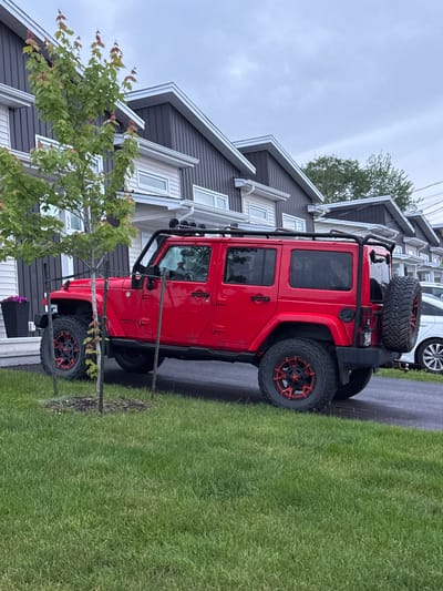 A red Jeep Wrangler is parked on asphalt in front of a row of modern townhouses with dark siding and white trim. The Jeep has a black roof rack, a spare tire mounted on the back, and black and red aftermarket wheels. A young tree with green leaves is planted in the grass between the Jeep and the townhouses. The sky is overcast and the grass is green and well-maintained. The scene is set in Dieppe, Canada.