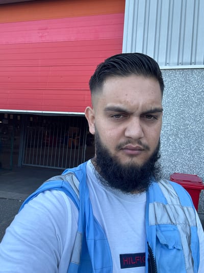 A man with dark hair styled in a fade and a full beard is taking a selfie outdoors. He is wearing a white t-shirt with a navy Hilfiger logo and a light blue and gray reflective vest. Behind him, a large red industrial roll-up door is partially open, revealing a dark interior with metal gates. To the right, a speckled gray building wall is visible, along with a red trash bin. The man's expression is neutral. The environment suggests an industrial or commercial area, potentially a warehouse or loading dock. The city of Boom, Belgium, is known for its industrial heritage and is often associated with port activities and large logistical centers, which aligns with the visual cues in the image.