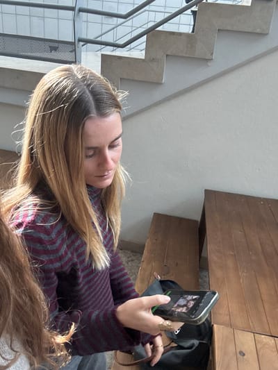 The scene is set indoors in Porto Salvo, Portugal. A female subject with long, straight, light brown hair, parted centrally, occupies the foreground, looking downwards and to her left. She wears a long-sleeved, high-necked sweater featuring horizontal stripes in dark red and grey. Her right hand, adorned with multiple rings, holds a black-cased smartphone horizontally; its illuminated screen displays an image containing at least two human faces. The top of another person's brown hair is visible in the bottom left corner. To the right of the primary subject, two dark brown wooden benches or tables are present, with a dark-colored bag resting near the smaller one. In the background, a modern staircase with light-colored steps and grey metal railings ascends against light-colored walls.