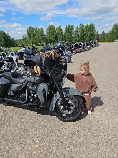 A young child in brown sweats stands beside a gray motorcycle in Borodinskogo Muzeya, Russia. The motorcycle is one of a row parked on a paved area. Trees and sky are in the background. A black motorcycle jacket is on the seat of the motorcycle.