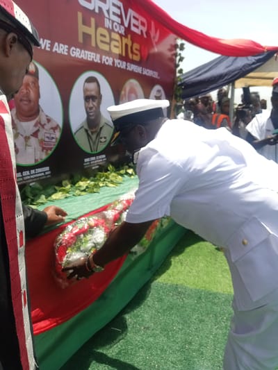 Here is a description of the image:
A funeral scene takes place outdoors. A banner reads "FOREVER IN OUR Hearts. WE ARE GRATEFUL FOR YOUR SUPREME SACRIFICE..." Photos of two uniformed men are displayed on the banner: Brig. Gen. DK Zirkushu and MHE. CHIEF CS APATA. The banner references "BATION HADIN KAI ATAGRAM" and the date "13TH NOVEMBER, 2021".
A uniformed man in white, possibly a naval officer, is laying a floral wreath on a coffin draped with a green and red cloth. Another man is standing beside the coffin. There are other people visible in the background, some holding cameras. The setting appears to be a solemn occasion, possibly a military or state funeral. The location is likely somewhere in Nigeria, based on the inscription "BATION HADIN KAI ATAGRAM". The image does not contain enough details to pinpoint the location in the city of Dilpurtu, Nigeria.