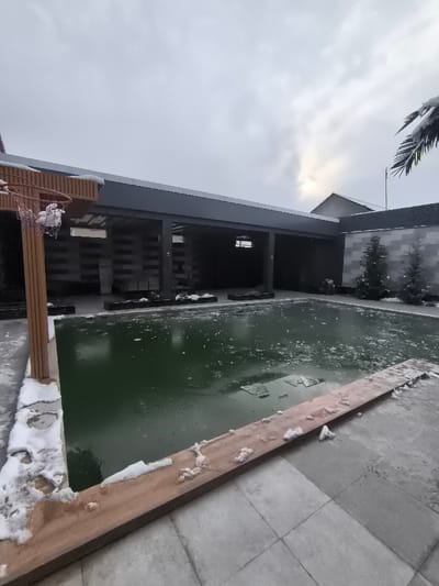 The scene depicts an outdoor residential or resort area in Charvak, Uzbekistan, under overcast winter conditions. A rectangular swimming pool, filled with dark green water, dominates the midground, its surface partially covered by sheets and fragments of ice. Snow patches accumulate on the light-colored tiled ground surrounding the pool and along a wooden-toned border. To the left, a wooden post supports a basketball hoop with a net, both partially dusted with snow. Behind the pool, a modern, open-sided structure with dark grey columns and a flat roof extends horizontally; its interior is shadowed. Further back, a light grey block-patterned wall is visible, accompanied by two small, snow-covered evergreen trees. A single snow-dusted palm frond extends into the upper right corner of the frame. The sky is largely grey and cloudy, with a faint bright spot of obscured sunlight in the upper right quadrant. No individuals or active interactions are present.