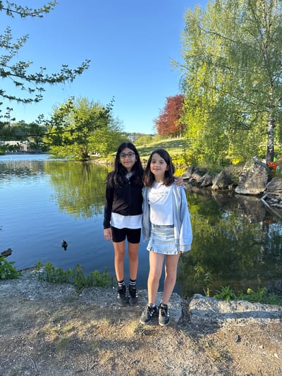Two young girls are captured in a bright, outdoor scene beside a calm body of water, likely a pond or lake, in Ulm, Germany. The image, rotated 90 degrees, shows the girls standing side-by-side, smiling towards the camera.
The girl on the left (when correctly oriented) wears a grey zip-up hoodie over a white t-shirt, a light blue denim skirt, and black sneakers with white socks. Faint text on her white t-shirt reads "Natural Sports Club." The girl on the right wears a black zip-up jacket over a white t-shirt, black shorts, black sneakers with striped socks, and glasses. Both girls have dark hair.
The setting is a natural, park-like environment with a clear blue sky. Lush green trees, some with light spring foliage and one with reddish leaves, line the water's edge and are reflected on its calm surface. Large, textured rocks form the bank where the girls are standing, and the ground is composed of dirt and small stones with some sparse greenery. A small bird or duckling can be seen swimming in the water near the girl in black.
The bright, natural lighting indicates a sunny day, possibly morning or late afternoon. The girls appear happy and relaxed, posing cheerfully for the photograph.