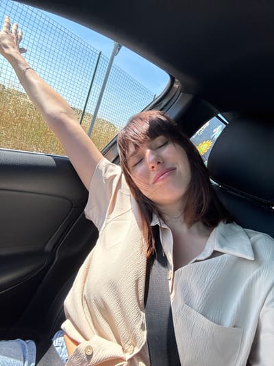 A woman is seated in a car, likely as a passenger. She is wearing a light-colored, button-down shirt and jeans. Her eyes are closed, and she has a relaxed expression. A seatbelt is across her chest. Her left arm is extended out the open car window. A fence and field are visible outside the window. The photograph may be from Nettuno, Italy.