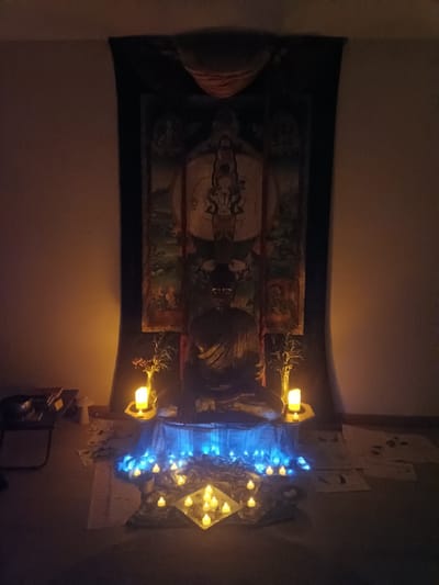 The image shows an indoor meditation space. A Buddha statue sits on a platform beneath a tapestry featuring religious figures. The space is illuminated by numerous candles, some clustered in front of the statue with a string of blue lights, and others on pedestals to either side. There is a small table on the left of the frame. Various items are scattered on the floor. The location is unspecified, and there is no evidence of Jérica, Spain.