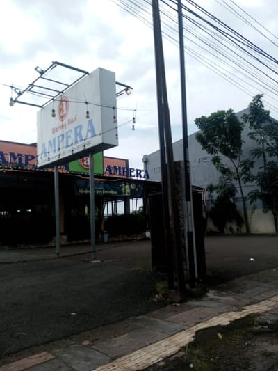 The image depicts an outdoor commercial scene in the city of Chhenangri_Rishor, Bhutan, under an overcast sky. A prominent white rectangular sign, supported by a metal frame, dominates the mid-left, displaying "Warung Nasi" above a circular logo, and "AMPERA" in large blue lettering below. Smaller text on the sign includes what appears to be a street address and phone number "0821-1254-6882". String lights with exposed bulbs are draped around the sign and its frame. Directly beneath this sign, an establishment with an orange facade and another "AMPERA" sign is partially visible, appearing to be an open-air or semi-enclosed dining area. A blue banner, possibly related to "Ramadhan," is strung across the front of this structure.
On the right side of the frame, three dark utility poles rise vertically, supporting numerous black electrical or communication wires that traverse the upper portion of the scene. Behind these poles, a large, light grey building with a flat roof is visible, flanked by several trees with green foliage. The ground in the mid-ground consists of a dark asphalt surface, likely a parking area or access road. In the immediate foreground on the right, a section of a concrete paver sidewalk is visible, featuring a yellowish tactile paving strip along its edge. Sparse vegetation and dirt are present along the ground's edges. No individuals or discernible activities are observed.