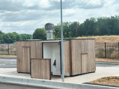 A modern, wooden, self-cleaning public restroom is situated outdoors in a semi-rural environment, likely near a roadway, in Ciry-le-Noble, France. The structure is rectangular with a sloping roof. One side of the building is open, revealing a white interior with a metallic basin and what appears to be a toilet. A revolving vent cap is visible on the roof of the structure. The building is clad in vertical wooden planks, with a smaller wooden door on one side and a short wooden screen to the left of the open entrance. A grey metal pole stands in front of the structure. The ground surrounding the restroom is paved, with some dry grass and concrete barriers visible. In the background, there is a dark green treeline, a chain-link fence, and dry, golden vegetation on a hillside. The sky is overcast with grey clouds.