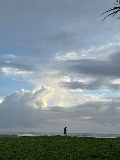 This media file captures a serene beach scene in Koggala, Sri Lanka, likely during the late afternoon or early evening. The sky is dramatically filled with a mixture of dark, imposing clouds and patches of bright, illuminating sky, suggesting dynamic weather conditions. A lone figure, appearing to be a person wearing a dark t-shirt and light-colored shorts, stands on the edge of a vibrant green, dense vegetation. This lush greenery borders the sandy shore where gentle waves of the ocean are visible. The composition, with the figure and vegetation on the right, and the expansive sky and sea on the left, creates a sense of vastness and tranquility. There are no visible text elements in the image.