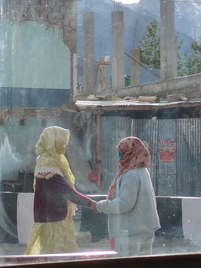In Padum, India, two women stand on a street. The woman on the left wears a yellow headscarf and a maroon sweater over a light yellow dress. The woman on the right wears a patterned headscarf and a light blue sweater and pants. They are holding hands and appear to be interacting. The background shows a damaged building with exposed brick and a corrugated metal roof. Construction materials, including concrete pillars and wooden beams, are visible. A sign with "S Z KING" printed on it is affixed to the corrugated metal. The scene appears to be viewed through a window, as evidenced by reflections on the glass.