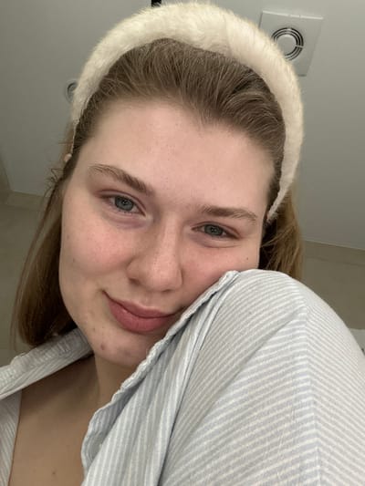 A young woman with fair skin, light brown hair, and gray eyes is shown in a close-up, tilted portrait. She wears a fuzzy white headband and a pale blue striped shirt, whose fabric is partially covering her right cheek. Her expression is gentle, with slightly parted lips. A ventilation fan is visible in the upper left corner of the image, suggesting an indoor setting.