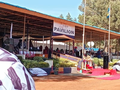 A pavilion marked "University of Eldoret PAVILION" with people seated under it. The structure has a blue and brown roof supported by metal posts. A flag is on the right. A stage area with red carpet and floral arrangements is visible in front. Landscaping with green shrubbery surrounds the stage. Seating and speakers are arranged on different levels under the pavilion.