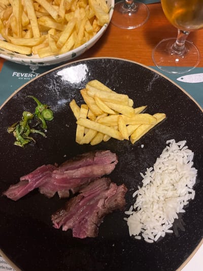 A black plate contains rice, french fries, fried green peppers, and sliced rare steak. A large bowl of french fries sits to the left. Two stemmed glasses, one containing a yellow beverage, are in the upper left. The plate's edge and a green and white tablecloth are also visible.
