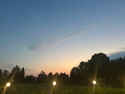 The image depicts a serene dusk scene in Ponteterra, Italy, characterized by a gradient sky transitioning from a deep blue at the zenith to warm hues of orange and pink near the horizon. A sliver of a crescent moon is visible in the upper left quadrant, accompanied by a faint star. Silhouetted against the colorful sky are the dark outlines of a variety of trees, some appearing mature and broad, while others are more slender and conical. In the foreground, a field of tall, dry grass stretches across the landscape. Several luminous, spherical garden lights are strategically placed, emitting a warm glow that illuminates the immediate surroundings. One light is prominent on the left, while two others are visible further to the right. In the distance, nestled amongst the trees, hints of buildings can be discerned, suggesting a residential or rural setting. The overall atmosphere is one of tranquility and natural beauty as day transitions into night.