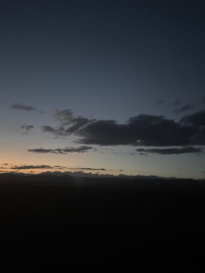 The image shows a dark, silhouetted landscape under a dusky sky. The lower portion of the image is almost entirely black, obscuring any details of the ground or any potential subjects. Above this dark expanse, a band of muted orange and yellow light suggests the last vestiges of sunset. This horizon is topped by a layer of dark, undulating clouds that obscure a range of mountains or hills. Higher up, scattered, darker clouds drift across a gradient of deep blue and gray sky. The overall impression is one of twilight settling over a vast, dark expanse, with the sky offering the only subtle hints of color. The location in Karnut, Armenia, is noted, but the image itself provides no specific visual cues to confirm or detail this location.
