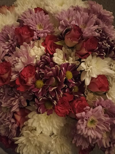 A close-up, overhead view of a bouquet of flowers. The bouquet contains roses in shades of red and pink, white chrysanthemums, and purple chrysanthemums with yellow centers. The flowers are densely packed and fill the entire frame. There is no visible background. The image is taken indoors. This image is not located in Annagry, Ireland.
