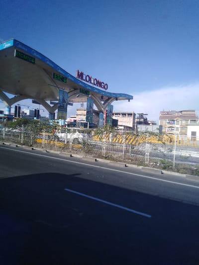 The image depicts a roadside view of a large, modern structure with the word "MLOLONGO" prominently displayed on top. The structure features a white, cantilevered roof with "Midea" branding. "ETC/MTC" signs are visible. A fence lined with vegetation and black and yellow striped barricades separates the structure from the road. Buildings are visible in the background against a clear, blue sky. The road surface is dark asphalt with a white line marking.