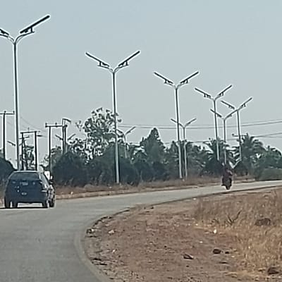 The image displays a paved road in Haifam, Nigeria, under a clear, bright sky. The road curves to the right, extending into the distance. A dark-colored sedan or hatchback is observed from the rear, traveling away from the viewer on the left side of the road. Further ahead, on the right side of the road, a motorcycle with a single rider is also moving away. Numerous tall, modern streetlights, each featuring a central pole with two upward-angled arms supporting light fixtures or solar panels, line both sides of the road. The road shoulders consist of dry, reddish-brown earth with sparse, dry vegetation. A dense line of green foliage, including several palm trees, forms a backdrop behind the streetlights. Older utility poles with visible overhead wires are present in the far-left background.