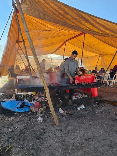 The image depicts an outdoor cooking and dining scene taking place under a large, temporary canopy. The setting appears to be a casual gathering or a makeshift eatery.

In the foreground, the ground is unpaved and consists of dirt and gravel, with scattered debris including pieces of paper, plastic, and a bright blue plastic bag. A long, light-colored wooden plank or branch lies diagonally across the lower right portion of the frame.

The central focus of the image is a long, rectangular cooking setup situated on the ground. Plumes of white smoke billow upwards from this setup, indicating an active fire or hot coals. A young man, dressed in a grey hooded sweatshirt and dark trousers, stands behind this cooking area, actively tending to the fire or food with a long tool, possibly a stirring paddle or poker. Several red buckets or containers are visible near the cooking pit. The ground around the cooking area is stained dark, suggesting prolonged exposure to fire.

Above and behind the cooking area, a large, expansive tent or canopy is erected. It is made of a coarse, orange-tan fabric, likely tarpaulin, supported by visible wooden poles and beams. The tent provides shade for a group of people seated at simple tables with white plastic chairs. These individuals are visible in the background, appearing to be eating or socializing under the diffused light of the canopy.

The scene takes place outdoors under a clear blue sky, visible in the upper left part of the image, suggesting good weather conditions during daytime, possibly morning or afternoon given the bright sunlight. No discernible text is visible in the image.