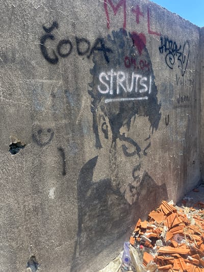 The image shows a concrete wall with graffiti in Gornji Kokoti, Montenegro. The wall is weathered and cracked, with several pieces of graffiti visible. In the foreground, there is a pile of broken terracotta roof tiles and some plastic bottles.

The graffiti on the wall includes:
* A portrait of a person in shadow, with the word "STRUTSI" written in white letters above their head.
* The word "Čoda" written in black letters on the left side of the wall.
* Other illegible graffiti tags and markings in black and red.
* A smiley face drawn in black on the left side of the wall.

The environment appears to be an outdoor, possibly abandoned or under construction, area. The sky is clear and blue. The overall impression is one of urban decay and artistic expression.