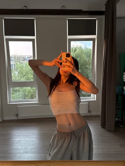 In a dimly lit room with wooden flooring, a young woman with long dark hair stands in front of a mirror, taking a selfie. She is wearing a white crop top and grey sweatpants. Her left arm is raised, holding a light yellow smartphone to capture the reflection. Sunlight streams in from two large windows, illuminating the scene with a warm glow. Outside the windows, a cityscape with trees and buildings is visible, hinting at the location in Zakharovo, Russia. A radiator is mounted on the wall beneath the windows. The room also contains a large grey curtain and some fitness equipment in the background.