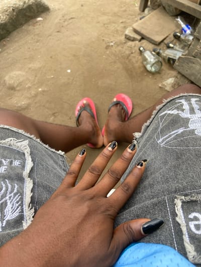 The image depicts a first-person view looking down at an individual's lap and lower extremities in Ojo, Nigeria. The subject's dark-skinned legs are visible, clad in gray denim shorts featuring frayed hems and white embroidered patterns. In the foreground, the subject's left hand is prominent, showing dark skin and fingernails painted with black polish that is significantly chipped and peeling, exposing the natural nail beneath. The thumb has a longer, pointed black nail. In the mid-ground, the subject's bare feet, also dark-skinned, are seen wearing pink flip-flops with black straps. The ground beneath the subject is light brown, appearing to be sandy or dusty soil. To the upper right, multiple empty glass bottles, at least five, are scattered on the ground near wooden planks and other debris. One bottle contains a residual blue liquid. A slight elevation of sandy earth is visible on the upper left.
