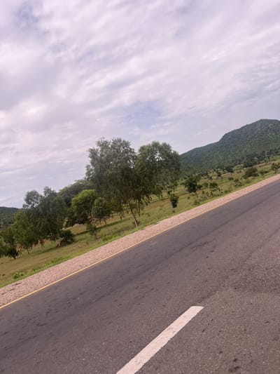 The image depicts a roadside scene in Minda, Nigeria, characterized by a paved road running alongside a green, hilly landscape dotted with trees. The perspective is from a low angle, looking diagonally across the road and into the landscape.
The road occupies the right side of the frame, its asphalt surface showing some wear and tear, with a white line marking the lane. A yellow line borders the road on the left. Beyond the road, a strip of gravel and then a grassy field with scattered trees stretches into the distance. In the background, a lush, green hill rises, covered in dense vegetation. The sky above is overcast with wispy clouds, suggesting a daytime scene, possibly in the late morning or afternoon.
The overall impression is one of a rural or semi-rural setting, with a sense of openness and natural beauty. There are no people or specific objects that indicate a particular event or activity. The environment appears tranquil, with no obvious signs of human activity beyond the road itself. The image captures the essence of the Nigerian landscape, with its vibrant greenery and rolling hills.