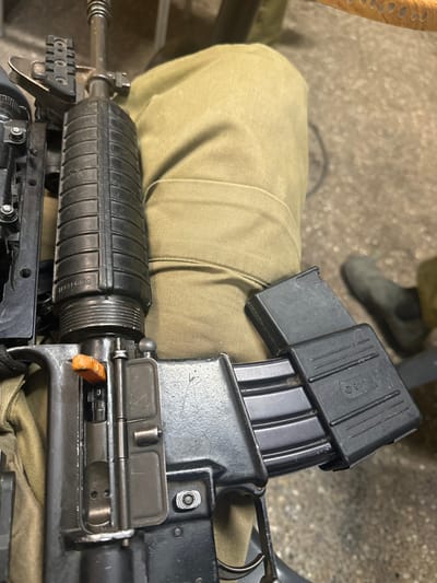 The image shows a black semi-automatic rifle resting on the lap of a person wearing khaki pants. The rifle is positioned vertically with the barrel pointing upwards. The upper receiver of the rifle is visible, and the charging handle is in the forward position. The rifle has a black handguard with a black magazine inserted. The magazine is closed with a black cover. The rifle is resting on the person's lap, and the bottom of the rifle is partially obscured by the person's clothing. The coordinates provided, latitude 31.86, longitude 34.811, indicate a location in Kiryat Ekron, Israel. The environment appears to be indoors, likely a residential or military setting. The image appears to have been taken with a mobile phone camera.
