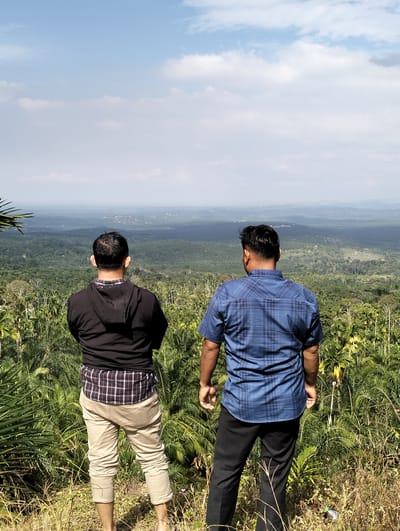 Two men stand with their backs to the camera, looking out over a lush, green landscape under a partly cloudy sky. The man on the left wears a black hooded jacket over a plaid shirt and light-colored pants. The man on the right is dressed in a blue plaid shirt and black pants. Both men appear to be in their late 20s or 30s. The foreground consists of dry grass and palm fronds. The background reveals a vast expanse of dense forest, with a hazy, mountainous horizon in the distance. The location is identified as Kilometer Delapan, Indonesia.