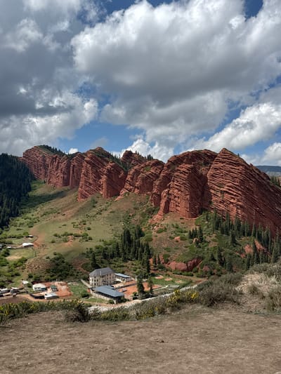 This media file captures a wide, scenic view of the Jeti-Ögüz gorge in Kyrgyzstan. The primary subjects are the striking red rock formations that stretch across the left and central parts of the image. These massive, layered cliffs, characteristic of Jeti-Ögüz, feature deep striations and varying shades of reddish-brown, contrasting with patches of dark green pine trees and other vegetation growing on their slopes and at their bases.
In the middle ground, nestled at the foot of these geological features and surrounded by green, grassy hills, is a collection of buildings. A prominent multi-story structure, possibly a hotel or resort, with a light-colored exterior and darker roofs, is visible. Several smaller buildings and outbuildings are also present, some resembling houses or support facilities. Towards the left of these structures, a few circular, manicured green areas suggest a golf course or recreational space. Further in the background, closer to the base of the red rocks, more distant, smaller structures or yurts can be observed.
The sky above is partially cloudy, showcasing a mix of bright blue patches and large, white to grey cumulus clouds. The foreground on the right consists of dry, earthy terrain, possibly a hillside or a dirt track, with patches of dry grass. A distinct line of small yellow flowers grows along what appears to be a fence or boundary separating the drier foreground from the greener developed area.
The image depicts a tranquil, daytime scene, likely under fair weather conditions, given the bright light and scattered clouds. No specific human activity is visible, but the presence of buildings indicates human settlement or a tourist destination within this rugged natural landscape. There is no visible text in the image.