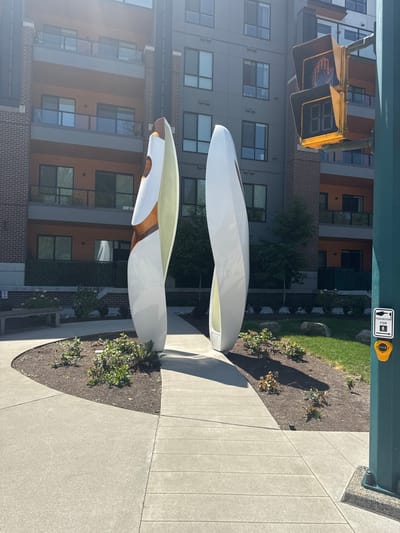 An outdoor scene in Port Moody, Canada, features a modern apartment building with balconies and large windows. In the foreground, two tall, white, abstract sculptures stand on a paved walkway. One sculpture has a copper-colored interior, while the other is entirely white. A traffic light with a pedestrian signal displaying a hand symbol and a countdown timer showing "88" is visible to the right of the sculptures. A yellow pedestrian crossing button is mounted on a green pole. Landscaping with bushes and grass surrounds the sculptures and the walkway. The sun casts shadows across the scene, indicating it is daytime.