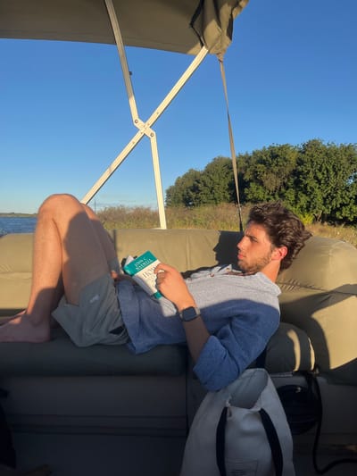 A man is sitting on a khaki-colored cushioned seat under the partial shade of a canvas roof supported by a white metal frame. The man has dark hair and a beard and is wearing a light blue patterned shirt, khaki shorts, and an Apple Watch. He is holding and reading a book titled "How Successful People Think" by John C. Maxwell. Behind him is a khaki-colored cushioned backrest. A tote bag and some other dark-colored items are partially visible behind the backrest. The background shows green trees and a blue sky. The bottom of the image shows a glimpse of water. Based on the context provided in the prompt, this scene is likely taking place in Victoria Falls, Zimbabwe.