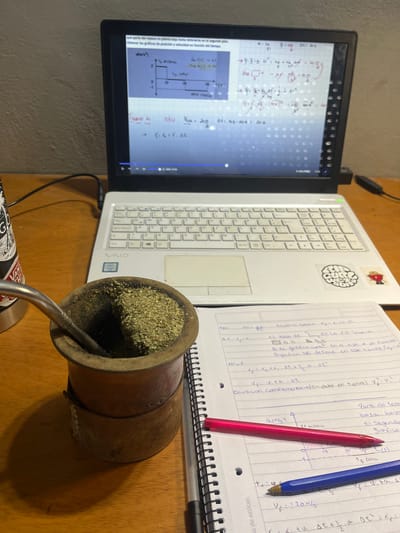 The image shows a desk setting with academic materials. A laptop displays equations and graphs, with handwritten notes beside it, including equations in Spanish. A gourd filled with yerba mate and a metal straw are in the foreground. A red and a blue pen lie on the notepad. The location in the city of Gerli, Argentina, is not indicated in the image.