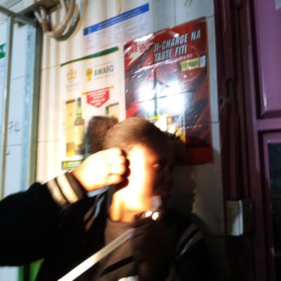 A medium indoor shot shows a blurred person with dark skin and hair, wearing a dark jacket with light-colored stripes on the sleeves. The person is holding up their fist in front of their face. In the background, there are posters on a wall, including an advertisement for "JI-CHARGE NA TASTE FITI." A bottle of alcohol is visible on another poster. The location is likely a shop or business in Kinoo ward, Kenya. There is a pinkish-purple door or frame to the right side. Cables are hanging at the upper part of the image.